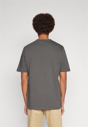 Mężczyzna o krótkich kręconych włosach, ubrany w gładki ciemnoszary t-shirt z krótkim rękawem i beżowe spodnie, stojący tyłem na białym tle.