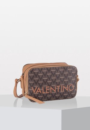 Prostokątna brązowa torebka typu crossbody Valentino z zamkiem błyskawicznym, ozdobiona powtarzającymi się logo Valentino i skórzanym paskiem, na białym postumencie.