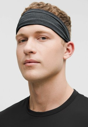 Ung mand med kort krøllet hår iført sort atletisk pandebånd og sort t-shirt med rund halsudskæring, ser let til siden.