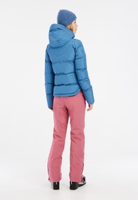 Hellblaues Pufferjacke mit Kapuze, mit gesteppten Abschnitten. Kombiniert mit pinken Skihosen und schwarzen Skischuhen, von hinten betrachtet.