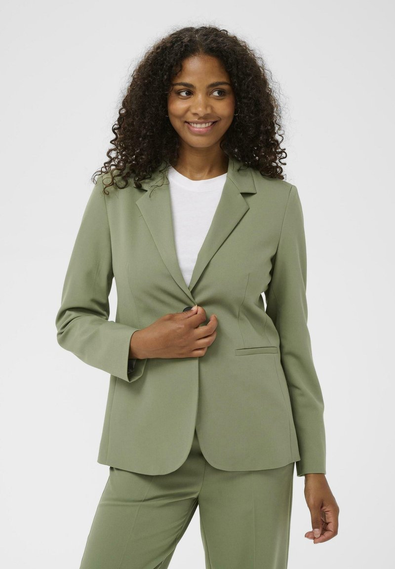 Blazer vert olive en tissu lisse, avec une fermeture à un bouton, des revers crantés et des poches latérales. Porté sur une chemise blanche.