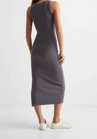 Donna che indossa un vestito midi fitted grigio scuro senza maniche e sneaker bianche, in piedi con la schiena rivolta alla macchina fotografica su uno sfondo neutro.