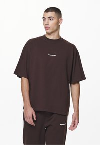 Barna, oversized pamut póló, kerek nyakkivágással és rövid ujjakkal. Az elején egy kis fehér logó található.