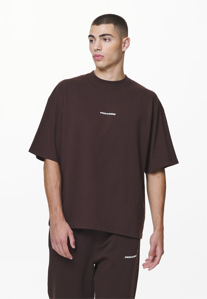 Barna, oversized pamut póló, kerek nyakkivágással és rövid ujjakkal. Az elején egy kis fehér logó található.