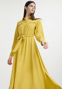 Abito camicia giallo con bottoni, maniche lunghe, due tasche sul petto, vita con cintura e uno spacco laterale, realizzato in un tessuto testurizzato.