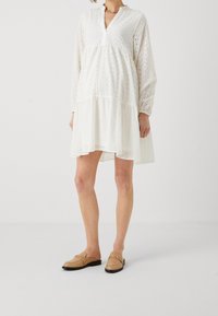 Weißes Augenmuster-Kleid mit langen Ärmeln, V-Ausschnitt, gestuften Rock und lockerem Schnitt. Kombiniert mit tanfarbenen Slippern mit einem metallischen Akzent.