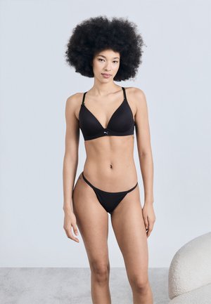 Μοναχική νεαρή γυναίκα με σγουρά μαλλιά, που φοράει μαύρο σουτιέν και εσώρουχα, στέκεται μπροστά σε ανοιχτό γκρι φόντο κοντά σε μια λευκή καμπυλωτή καρέκλα.