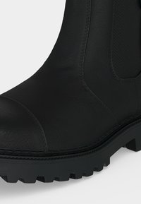 Botte en cuir noir avec bout arrondi, panneau latéral élastique à motif chevron, et semelle épaisse avec crampons profonds pour une bonne accroche.
