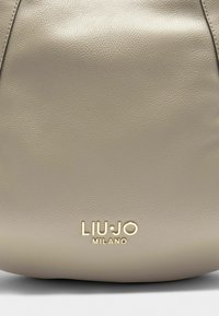 Borsa a mano in pelle beige con una texture a pietra. Presenta un logo del brand in oro in rilievo "LIU•JO MILANO" nella parte inferiore. Forma arrotondata.