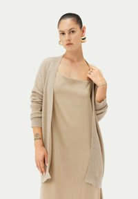 Beige stickad cardigan med mönstrad textur, öppen framsida och långa ärmar, bärs över en slät beige klänning. Enkel, neutral färgpalett.