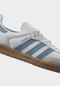 White and light blue sneaker with three blue stripes, suede accents, and a textured brown rubber sole. Features "Samba" branding.