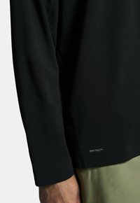 Mustat pitkät hiharat urheilupaita, joka on valmistettu kosteutta siirtävästä kankaasta, jossa on "DRYTEC™" -tagi helman lähellä ja sileä pinta.