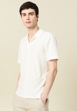 Junger Mann mit kurzen dunklen Haaren, trägt ein weißes, strukturiertes Polohemd mit kurzen Ärmeln und helle beige Hose, Hände in den Taschen, neutraler Hintergrund.