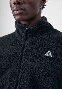 Fleece bunda v čiernej farbe s textúrovanou tkaninou, vysokým golierom, zipsom a malou trojuholníkovou logom. Má mäkký a teplý dizajn.