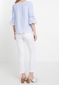 Woman wearing light blue off-shoulder blouse with eyelet lace trim, white slim-fit pants, and white heeled sandals, standing against white background.