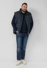 Granatowa kamizelka puchowa na ciemnym swetrze, niebieskich dżinsach i kremowych sneakersach. Kamizelka ma wysoki kołnierz i zapięcia na napy.