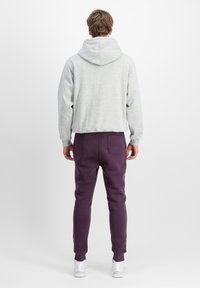 Grau hoodie mit einer großen Kapuze mit Kordelzug; kombiniert mit dunkelvioletten Jogginghosen mit einer Gesäßtasche. Model steht mit dem Rücken zur Kamera.