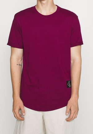Mann trägt ein schlichtes magentafarbenes T-Shirt mit kurzen Ärmeln und kleinem schwarzem Calvin Klein-Logo-Patch am Saum, kombiniert mit hellen Shorts.