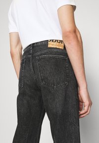 Persona que lleva un pantalón de mezclilla negro y una camisa blanca de manga corta, mostrando el bolsillo trasero y una etiqueta de cuero marrón en la cinturilla.