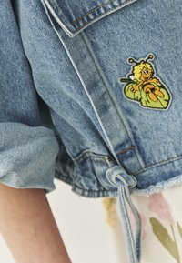 Denimjack met opgerolde mouwen, voorzien van een kleurrijke geborduurde patch van een cartoonbij aan de voorkant. Verstelbare strik in de taille.