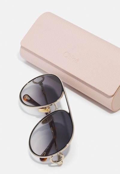 Óculos de sol com lentes cinza-escuras e armação em metal dourado. Inclui uma caixa de pele texturizada rosa-claro com um logótipo em relevo.