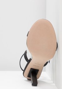 Sålen på en højhælet sandal med en firkantet sort hæl, sorte og hvide stropper, beige sål og "HUGO Made in Italy 37" præget.