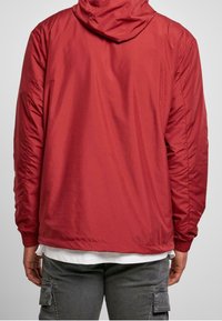 Mężczyzna w czerwonej kurtce z kapturem, białej koszuli pod spodem i ciemnoszarych spodniach cargo, widziany od tyłu na jednolitym tle.