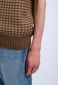 Trui in bruin en beige breiwerk met een gestructureerd patroon, geribde zoom en een genaaide logo label. Gedragen over blauwe spijkerbroeken, wat de handdetail laat zien.