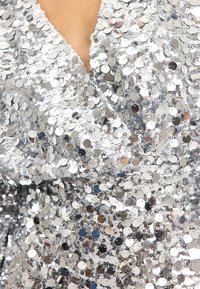 Tela cubierta de lentejuelas plateadas con un escote en V profundo, que exhibe lentejuelas circulares que reflejan la luz, creando un efecto texturizado y brillante.