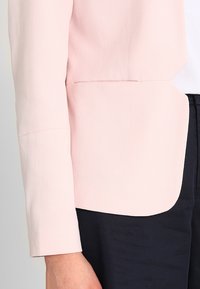 Světle růžový blazer z hladkého materiálu, s přiléhavým střihem, dlouhými rukávy a zaobleným spodním okrajem. Nosený přes tmavé kalhoty.