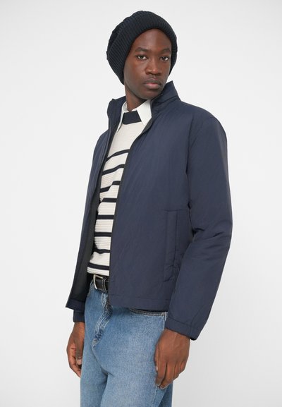 Nαυτικό puffer μπουφάν με ψηλό γιακά, φερμουάρ και τσέπες στα πλάγια. Φορεμένο πάνω από ένα ριγέ πουλόβερ και ανοιχτό μπλε τζιν παντελόνι, με ένα πλεκτό σκουφάκι.