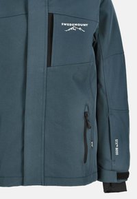 Niebieska kurtka odporna na warunki atmosferyczne z czarnymi akcentami, wieloma kieszeniami, zamkiem błyskawicznym z przodu i detalami z logo. Stworzona z myślą o aktywnościach na świeżym powietrzu.