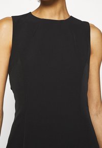 Robe noire sans manches avec un col montant, coupe ajustée, légères épaulettes et texture lisse. Aucune motif ou élément métallique visible.