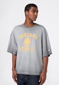 Junger Mann in einem grauen oversized T-Shirt mit gelbem "Indiana State"-Text und Emblem, schaut nach rechts vor einem weißen Hintergrund.