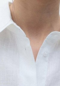 Close-up van de nek en bovenste borstkas van een persoon die een witte blouse met open kraag draagt.