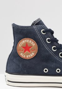 Námořnické nubukové vysoké tenisky s kulatou hnědou nášivkou, na které je červená hvězda a text "Converse All Star"; bíl�á gumová podrážka s černými pruhy.