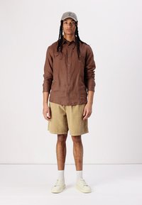 Chemise à manches longues marron, short en velours côtelé beige, baskets blanches, casquette grise, debout contre un fond uni.