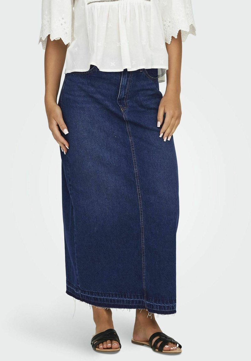 Jeansmaxikjol i mörkblå, med rak skärning, framstygn och fransig fåll. Kombinerad med en vit blus och svarta sandaler.