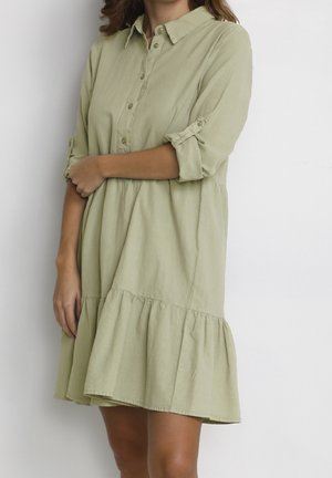 Femme portant une robe boutonnée vert clair avec des manches retroussées et un ourlet à volants froncés, debout sur un fond blanc uni.