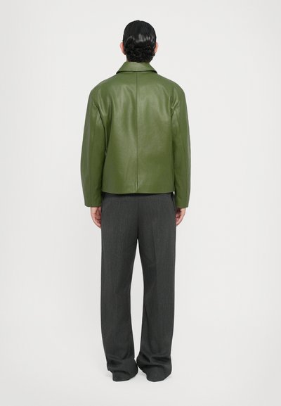 Chaqueta de cuero verde con un collar clásico, corte relajado y textura suave. Combinada con pantalones gris sastre, mostrada desde la parte de atrás.