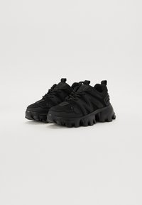 Chaussures de sport noires avec un dessus en maille et en synthétique, semelle épaisse avec des crampons prononcés, et un design multicouche pour une texture ajoutée.