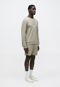 Férfi bézs színű hosszú ujjú ingben és hozzá illő rövidnadrágban, fehér zokniban és sportcipőben, egyszerű fehér háttér előtt áll.