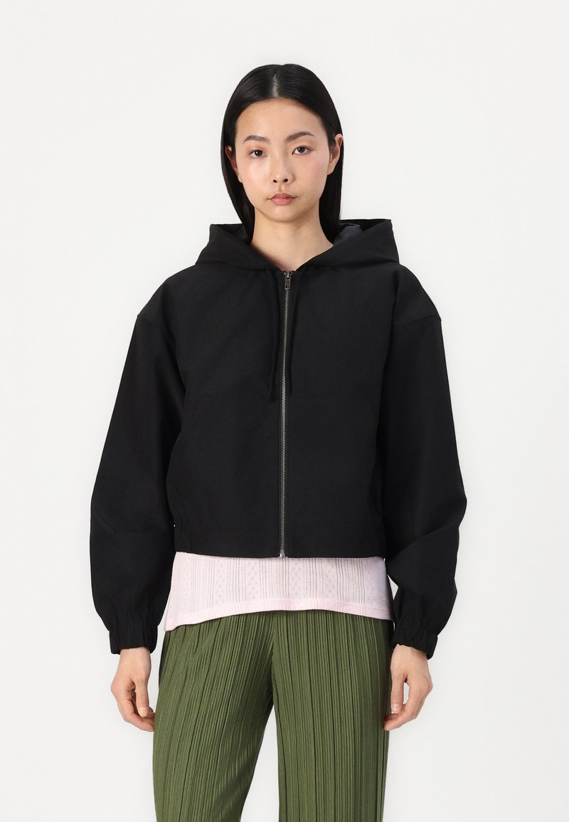 Black cropped zip-up jacket with a hood, featuring wide sleeves and a smooth texture. Worn over a light pink top and green pleated pants.