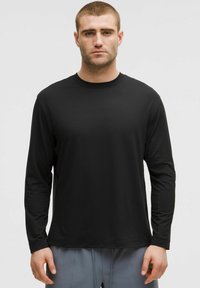 T-shirt nera a maniche lunghe in tessuto morbido, con colletto rotondo e vestibilità rilassata. Indossata con pantaloni grigi chiari.