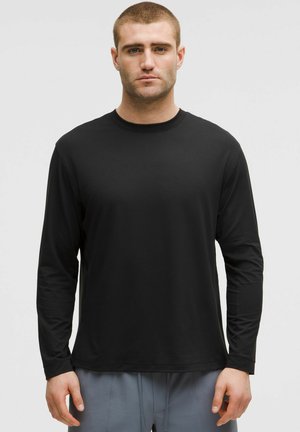 Czarna, długą rękaw t-shirt z gładkiego materiału, z okrągłym dekoltem i luźnym krojem. Noszona z jasnoszarymi spodniami.