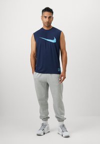 Ærmeløs marineblå Nike shirt med et turkist logo, parret med lysegrå sweatpants og hvide sneakers. Afslappet atletisk stil.