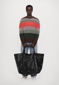 Black leather tote bag with silver studs, large shape, soft texture, and dual handles. Model wearing a multicolored striped sweater and jeans.