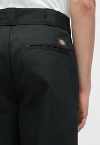 Sorte bukser med en glat overflade, der har en baglomme og et Dickies logo-patch. Designet inkluderer en lige pasform og en talje.