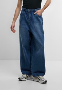 Jeans in denim blu con gamba larga e vita alta, con tasche anteriori e una finitura lavata, abbinati a sneaker sportive grigie e argentate.