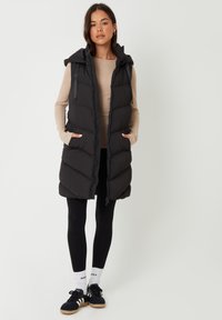 Frau trägt eine lange schwarze gesteppte Weste über einem beigen Langarmshirt, schwarze Leggings, weiße Socken und schwarze Turnschuhe, steht vor einem weißen Hintergrund.